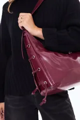 Cartera tipo hobo de material sintético con acabado brillante, cierre superior con cremallera, correa ajustable y laterales con cordones decorativos.