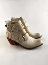 Bota corta de cuero natural color beige, con taco de madera, puntera afinada y cierre trasero. Cuenta con tiras con hebillas metálicas y detalles pintados a mano en la parte trasera.