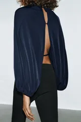 Top azul marino de tejido fluido con cuello redondo y manga larga abullonada con aberturas. Presenta una abertura en la espalda con tira del mismo tejido y cierre de ganchos metálicos.