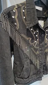 Campera de jean negra, corte a la cintura, con bordados decorativos, tachas plateadas y flecos de cadenas en la parte frontal y en las mangas.