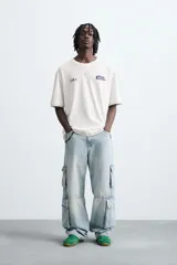 Camiseta de manga corta y cuello redondo, color blanco roto, con efecto de doble capa (la capa inferior gris asoma en el ruedo). Presenta estampados pequeños en azul marino en el pecho: un logo con texto 'Idle Eden' y otros símbolos.