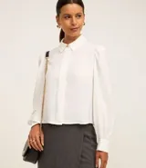 Camisa blanca de crepé con cuello inglés, manga larga abullonada con puños y abotonado frontal. Presenta un botón dorado diferenciado en el cuello.