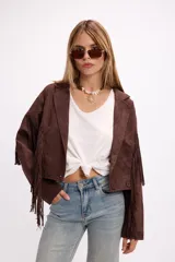 Chaqueta de efecto gamuza en color tostado, con solapas amplias, cierre frontal con un botón y flecos largos decorativos en la parte frontal y trasera de las mangas. Presenta una estructura definida y forro interior.