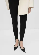 Leggings negros de corte entallado con estribos en la parte inferior, confeccionados en tejido elástico con costuras verticales decorativas.