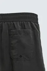 Bañador de hombre negro, largo hasta la rodilla, con acabado aterciopelado. Presenta cintura elástica ajustable con cordón, bolsillos laterales y un bolsillo trasero de plastrón. Incluye forro interior.