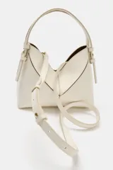 Bolso tipo mini shopper de diseño estructurado en color beige, con forma de V en la parte superior y asa de mano fija. Presenta costuras visibles y detalles metálicos en los laterales.