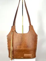 Bolso matero de cuero color marrón con textura tipo piel de cocodrilo en la parte superior y cuero liso en la parte inferior. Tiene dos asas de hombro, cierre superior y detalle de flecos de cuero en un lateral.
