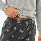 Conjunto de pijama de dos piezas confeccionado en tejido polar. Incluye una sudadera gris de cuello redondo con detalle bordado en el pecho y un pantalón largo de color gris oscuro con estampado de letras y puños elásticos en los tobillos.