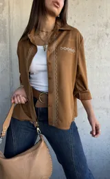 Camisa de gamuza color camel, con cuello clásico, cierre frontal de botones y detalle de bordado geométrico en color crema a lo largo de la botonadura y en el bolsillo frontal. Presenta mangas largas con puños doblados.