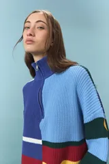 Sweater de tejido de punto grueso con diseño de bloques de color y rayas horizontales en tonos azul, rojo, amarillo y verde. Presenta cuello alto con cierre frontal parcial y mangas largas con puños ajustados.