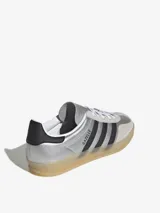 Zapatillas Adidas Gazelle Indoor plateadas con las tres tiras laterales en negro, contrafuerte trasero en negro y suela de goma color caramelo.