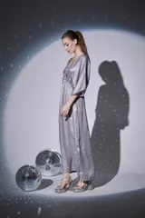 Vestido largo de fiesta en color plateado con acabado brillante. Presenta escote en V cruzado, mangas tipo kimono 3/4 y cintura elástica con frunces que dan caída fluida a la falda.