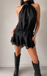 Vestido corto negro con escote halter ajustable con lazo en el cuello y espalda abierta con tira regulable. Presenta cintura fruncida con efecto fajín y falda corta con volados. El tejido es satinado con textura de rayas sutiles.