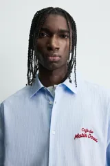 Camisa de algodón de corte regular, color celeste con finas rayas blancas verticales. Presenta un bordado en color rojo en el pecho con la frase "Café du Matin Doux".