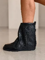 Bota corta de cuero gamuzado color marrón, con diseño de polaina superpuesta y detalle de hebillas metálicas en la parte posterior del talón. Cuenta con suela plana de goma negra y horma amplia.