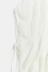 Top blanco de cuello redondo y mangas tipo capa, con textura fruncida, bajo asimétrico, lazos decorativos en el escote y en los laterales, y cierre posterior con botón.