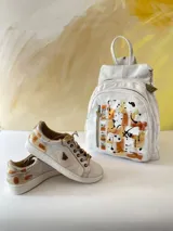 Mochila de cuero blanca con diseño artístico pintado a mano en tonos dorados, marrones y negros en el frente, múltiples bolsillos con cierre y herrajes de metal.