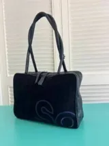 Cartera de mano tipo bolso, color azul oscuro, con diseño rectangular y asas largas trenzadas. Presenta un detalle de logo bordado en tono azul más claro en la parte inferior frontal.