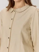 Camisa de denim con silueta levemente entallada, cuello bebé y mangas largas con sutil frunce en los hombros. Presenta costuras curvas decorativas con pespuntes en contraste y cierre frontal con botones.