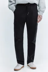 Pantalón cargo negro de corte slim fit, con cintura elástica y cordón ajustable. Presenta bolsillos al bies, bolsillos cargo en las piernas con solapa y botones a presión ocultos, y bolsillos traseros con solapa. Las piernas son ajustadas con costuras y pinzas en las rodillas.