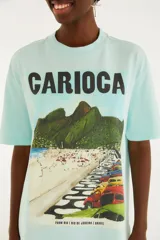 Remera celeste de corte holgado, con cuello redondo, mangas cortas y un gran estampado frontal que dice "CARIOCA" junto a una ilustración de playa y autos.