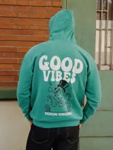 Canguro de algodón peletizado en color verde, con capucha y cordones ajustables. Presenta un bolsillo frontal tipo canguro y un estampado gráfico en el pecho con la frase 'Good Vibes' y un personaje animado.