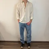 Camisa de manga larga color beige claro, con cuello clásico, cierre frontal con botones y logo bordado en el pecho. Corte regular.