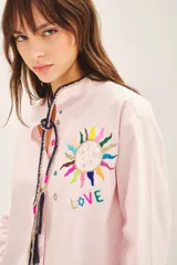 Camisa de algodón color rosado con cuello mao, cierre frontal con botones de nácar y bordado artesanal multicolor en el pecho. Presenta un cordón decorativo en el cuello con borlas.