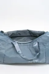 Bolso de viaje gris con doble asa corta y cierre superior. Estampado frontal con la frase "The Tote Bag" en letras blancas.
