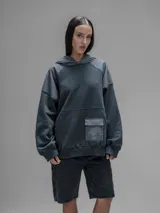 Hoodie oversize de color gris oscuro, confeccionado en frisa de algodón con recortes en textiles técnicos. Presenta capucha forrada, bolsillo frontal tipo canguro y un bolsillo adicional con solapa en la parte inferior.