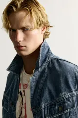 Campera de denim de algodón en color azul, con lavado efecto desgastado, cuello solapa y cierre frontal de botonadura metálica. Presenta bolsillos con solapa en el pecho, mangas largas con puños abotonados y trabilla ajustable en la cintura posterior.