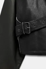 Chaqueta corta de piel 100% color negro, de estilo cruzado con cierre de hebilla lateral. Cuenta con cuello solapa ancho, manga larga con puños estructurados y forro interior.
