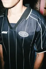 Camiseta de estilo deportivo con cuello polo, confeccionada en tejido técnico negro con finas rayas verticales blancas. Presenta paneles laterales en gris, mangas cortas con bordes en contraste y detalles gráficos estampados en el pecho.