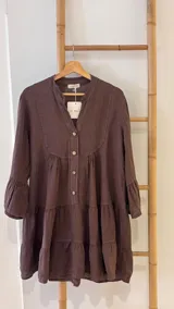 Vestido corto de lino color marrón oscuro, con cuello mao y abertura frontal con botones de coco. Presenta un diseño amplio con frunces horizontales que crean volados en el cuerpo y mangas tres cuartos con terminación acampanada.