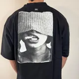 Remera negra de algodón pesado, corte oversize, con un gran estampado en la espalda en blanco y negro que muestra la parte inferior del rostro de una persona con un gorro tejido cubriendo los ojos y un cigarrillo en la boca.