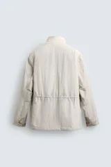 Cazadora técnica de corte relaxed fit, color beige, con cuello subido, manga larga y acolchado interior. Cuenta con múltiples bolsillos frontales con solapa, cintura ajustable con cordón interior y cierre frontal de cremallera oculta por solapa con botones a presión.