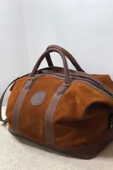 Bolso de viaje tipo duffel de cuero y gamuza color marrón, con asas de mano reforzadas y correa ajustable para el hombro. Presenta detalles de tiras de cuero liso en contraste, base estructurada y un parche circular con logotipo grabado en el frente.