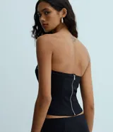 Blusa negra sin breteles, ajustada al cuerpo, con ojales metálicos distribuidos en la parte delantera y cierre de cremallera en la espalda. Confeccionada en bengalina con viscosa certificada.