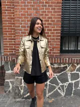 Chaqueta dorada metalizada con cuello camisero, cierre frontal con botones y bolsillos de parche en el pecho.