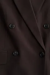 Blazer de color marrón oscuro, de corte ligeramente largo, con solapas de muesca y cierre de doble botonadura con botones oscuros. Presenta hombreras y bolsillos delanteros ribeteados con solapa.