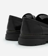 Zapato masculino tipo mocasín de color negro, confeccionado en poliuretano con efecto vinilo. Presenta punta redonda, detalle de antifaz y suela track gruesa con pespuntes en contraste.