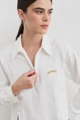 Remera de manga larga color blanco con cuello polo y cierre frontal parcial. Presenta costuras a la vista en contraste y un pequeño bordado en el pecho. El diseño incluye una abertura lateral en el ruedo.