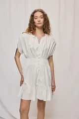 Vestido corto de algodón color blanco, con escote en V y botonadura frontal. Presenta mangas cortas holgadas, cintura ajustada con frunces y falda acampanada con terminación de volado en el ruedo.