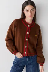 Cardigan de lana color marrón con detalles de bordes en color rojo. Presenta cierre frontal con botones, mangas largas con puños ajustados y un pequeño bordado decorativo en el pecho.