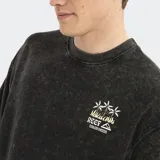 Remera de corte oversized en color negro con efecto lavado, cuello redondo y mangas cortas. Presenta un pequeño estampado gráfico en el pecho con el logo de la marca y palmeras.