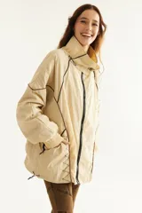 Campera rompevientos beige de nylon con diseño oversize, cuello alto, cierre frontal negro y detalles de costuras y ribetes en contraste.