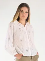 Blusa blanca de broderie con cuello camisero, cierre frontal con botones y mangas largas abullonadas con puños ajustados.