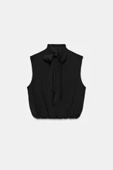 Blusa sin mangas blanca con estampado de lunares negros, cuello alto drapeado y nudo frontal en el bajo.