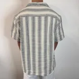 Camisa de manga corta con corte oversize, cuello tipo cubano y cierre frontal con botones. Presenta un diseño de rayas verticales en tonos crudo y gris.