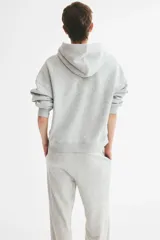 Campera canguro de felpa color gris melange, de corte holgado con interior cepillado suave. Cuenta con hombros caídos, capucha forrada, cierre frontal y bolsillos delanteros abiertos, con puños y bajo en punto acanalado.